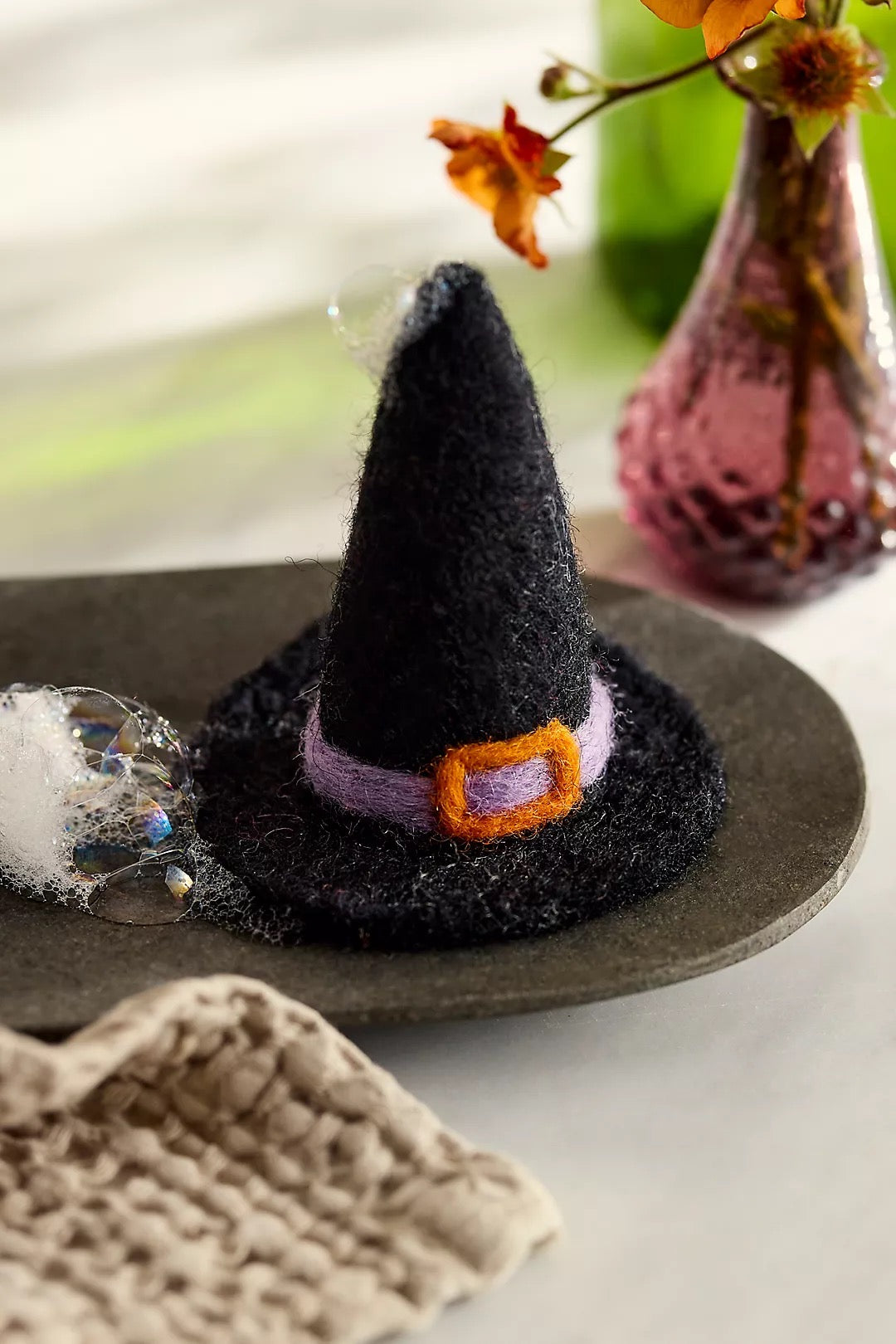 Black witch hat felted soap with a purple band and orange buckle on a dark surface, with a blurred background.