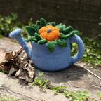 A handmade felt watering can decoration with a blue body, green leaves, and an orange pumpkin on top, placed on a weathered wooden surface outdoors.
