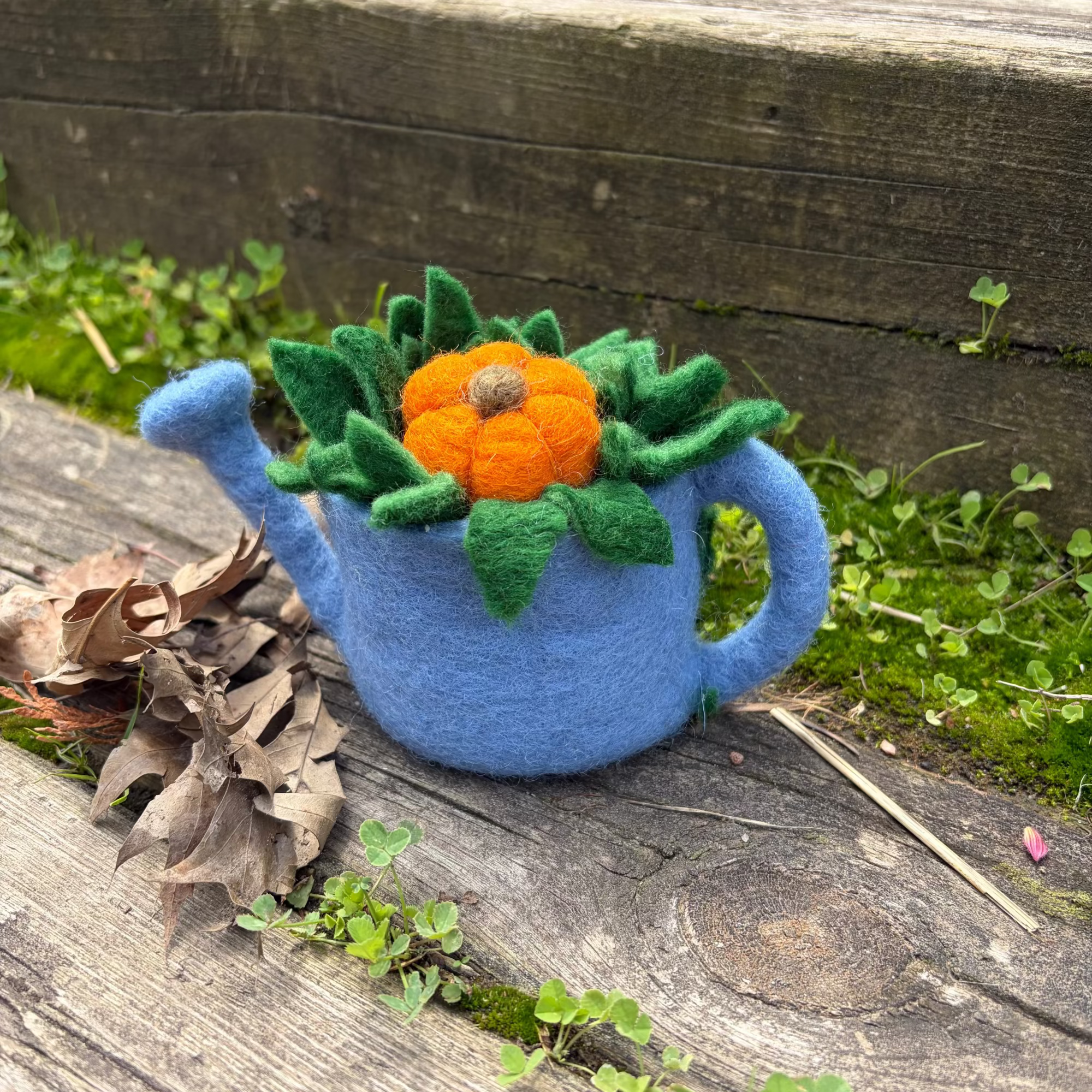 A blue felt watering can decoration with a green leafy top and an orange felt pumpkin, placed on a weathered wooden surface outdoors with grass and leaves around it.