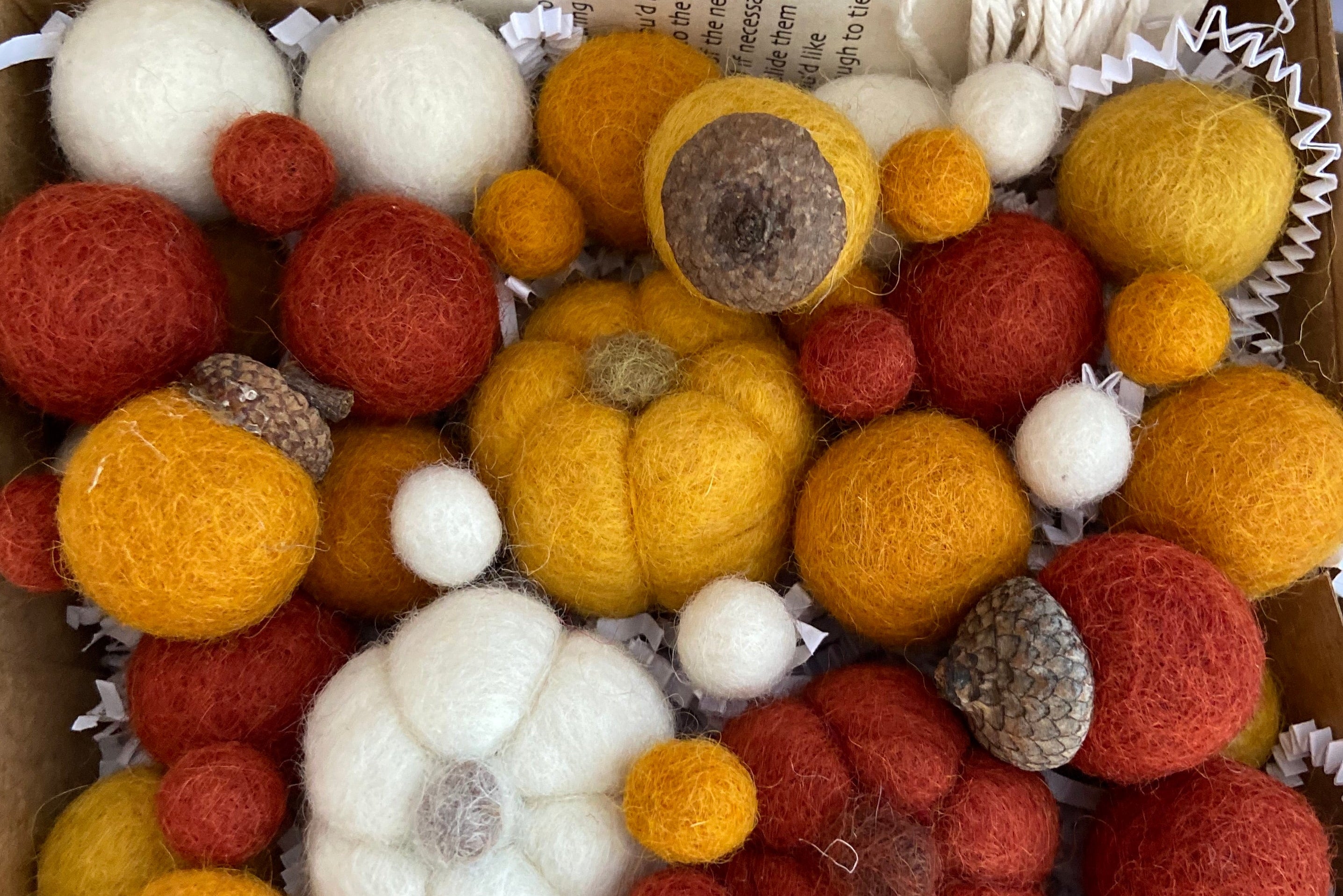 A collection of autumn-themed felted wool decorations, including pumpkins, acorns, and pom-poms in orange, yellow, red, and white, arranged in a cardboard gift box with white paper filler.