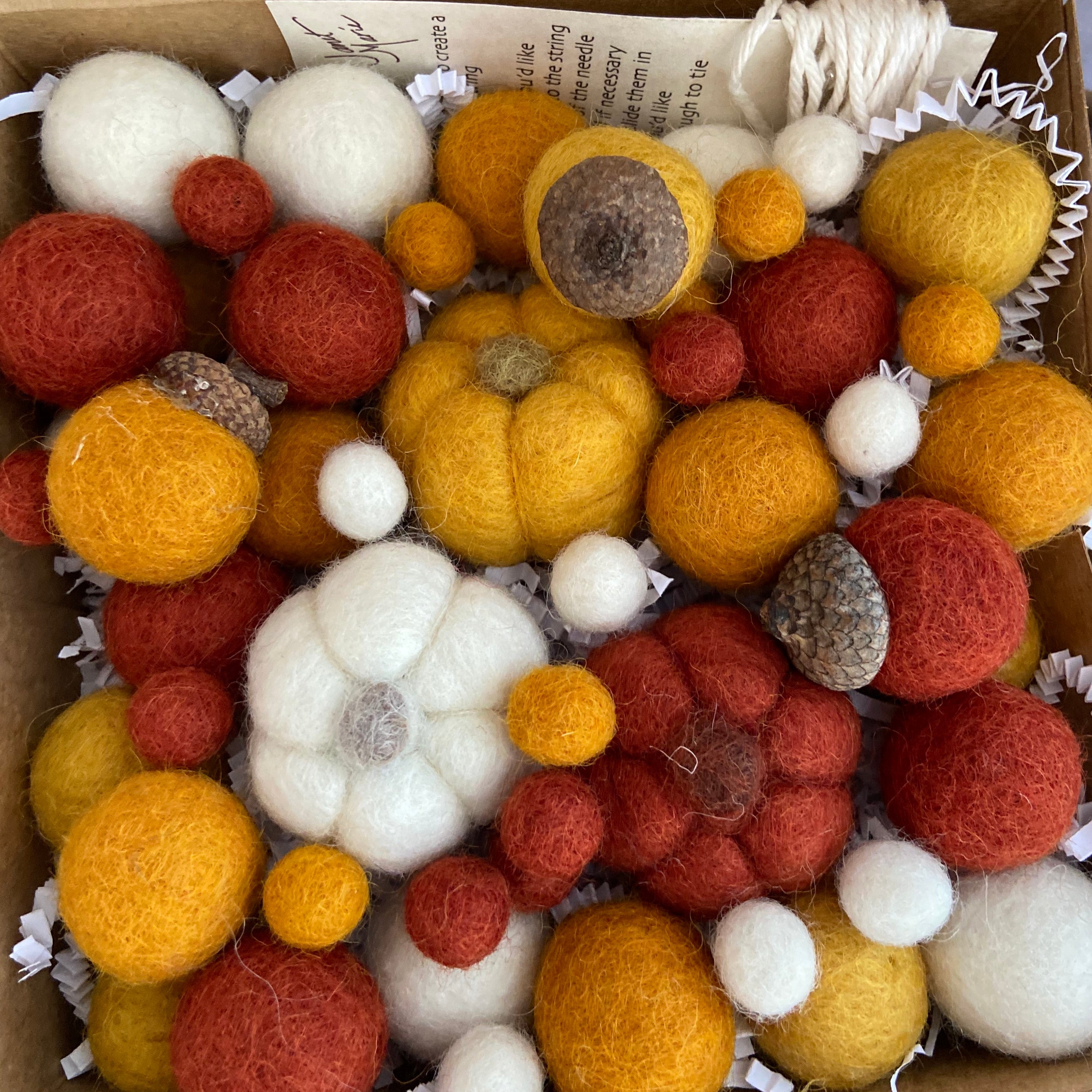 A close-up of autumn-themed felt and acorn decorations arranged in white paper packing material. The set includes round felt balls in mustard yellow, rust red, and cream, a textured felt pumpkin in mustard yellow, and two acorns with brown caps and gray-brown textured bases.